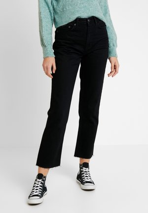 Personne portant un jean noir droits coupé court, des baskets montantes noires avec des lacets blancs et un pull en tricot bleu clair.