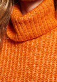 Pomarańczowy sweter z golfem wykonany na drutach, z prążkowaną teksturą, z wysokim kołnierzem i ciepłym, miękkim materiałem.