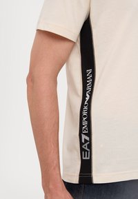 Béžové tričko s krátkým rukávem, které má černý boční panel s bílým logem "EA7 Emporio Armani" a kontrastním prošíváním podél okraje.