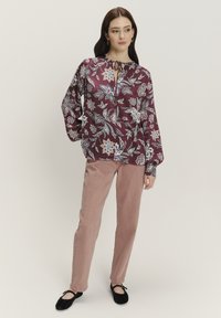 Bordeauxfarbene Bluse mit floralem Muster, langen Ärmeln und einem Bindeband am Hals; kombiniert mit hellrosa Cordhosen. Schwarze flache Schuhe runden das Outfit ab.