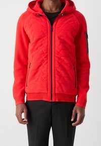 Man med en ljus röd vadderad zip-up hoodie med svarta dragkedjedetaljer och svarta byxor, stående med avslappnade armar.