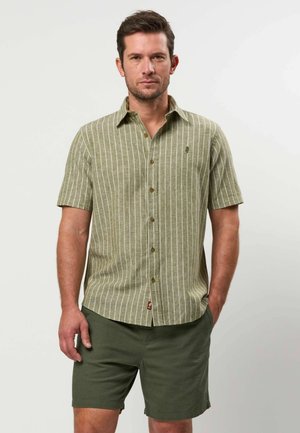 Man draagt een olijfgroen overhemd met korte mouwen en witte verticale strepen, gecombineerd met effen olijfgroene korte broek, staand met één hand in de zak.