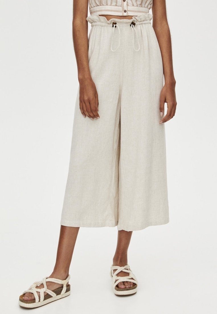 Pantalons en lin beige à jambes larges et taille à cordon. Texture lisse et longueur courte. Assortis à des sandales tressées à lanières.