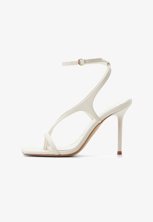 Sandalia blanca de tacón alto con tiras finas, un lazo para el dedo y una hebilla en el tobillo sobre un tacón de aguja delgado, sobre un fondo blanco.