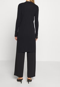 Langes schwarzes geripptes Kleid mit eng anliegenden Ärmeln, seitlichen Schlitzen und goldenen Knotendetails an den Manschetten, kombiniert mit weit geschnittenen, schwarzen Hosen und hohen Schuhen.