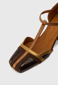 Chaussure en cuir marron avec un bout carré, présentant une combinaison de textures lisses et en daim, accentuée par des bandoulières dorées et un intérieur beige.