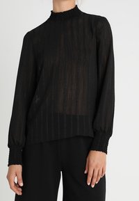Black, sheer blouse with vertical stripes, high smocked collar, and long balloon sleeves with smocked cuffs. Light texture, elegant design.