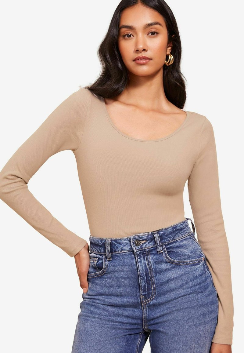 Beige hosszú ujjú bodysuit bordázott textúrával és kereknyakú fazonnal, világoskék, magas derekú farmernadrággal, elülső zsebekkel párosítva.