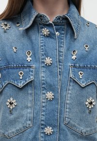 Τζιν πουκάμισο με μεταλλικά κουμπιά πατιλέτας διακοσμημένα με μαύρες και διάφανες πέτρες σε σχήμα λουλουδιού και σταγόνας, εμφανίζοντας γιακά και τσέπες στο στήθος.