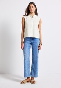 Ljusbeige ärmlös topp med mandarin-krage, knäppning fram och volangkanter, tillsammans med ljusblå vida jeans och beige sandaler.