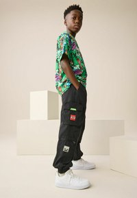 Zöld grafikai póló, amelyen rajzfilm motívumok találhatók, fekete cargo nadrággal párosítva, amely több zsebet is tartalmaz, és egy piros-fehér "TNT" címke díszíti. Fehér tornacipő.