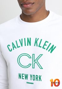 Witte sweatshirt met groene opdruk, met "CALVIN KLEIN" boven "CK" en "NEW YORK." Ronde hals en zachte textuur. Numeriek accent aan de onderkant.
