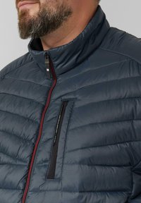 Marineblauer Steppjacke mit quiltierter Textur, hohem Kragen und rohem Reißverschluss. Verfügt über eine kleine Reißverschlusstasche auf der linken Brustseite.
