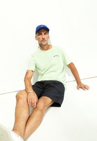 Lichtgroene katoenen t-shirt met logo, gecombineerd met donkerblauwe shorts. Zittende positie tegen een witte achtergrond, met een blauwe pet op.