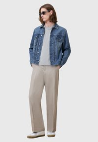 Giacca di jeans di un blu chiaro con due tasche sul petto, indossata sopra una camicia grigia. I pantaloni beige e le sneakers bianche completano l'outfit.