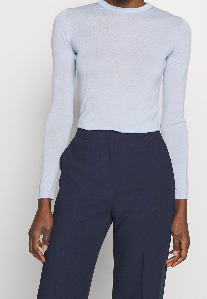 Femme portant un haut moulant bleu clair à manches longues, rentré dans un pantalon taille haute bleu marine sur mesure, se tenant devant un fond uni.
