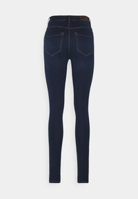 Jean skinny bleu foncé vu de dos avec deux poches arrière, passants de ceinture et coutures visibles sur un fond uni.