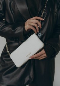 Portefeuille blanc texturé avec un logo doré, fermeture zippée et une bandoulière amovible, tenu dans une main. L'arrière-plan présente une veste noire.