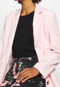 Světle růžový blazer s klopami, vyrobený z lehké tkaniny a s dlouhými rukávy, nošený přes černé tričko a sukni s květinovým vzorem.