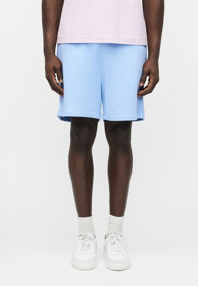 Inimene, kes kannab helerohelisi lühikesi pükse, heleroosat särke, valgeid sokke ja valgeid vabaajajalatsid, seisab valge ühetoonilise tausta ees.