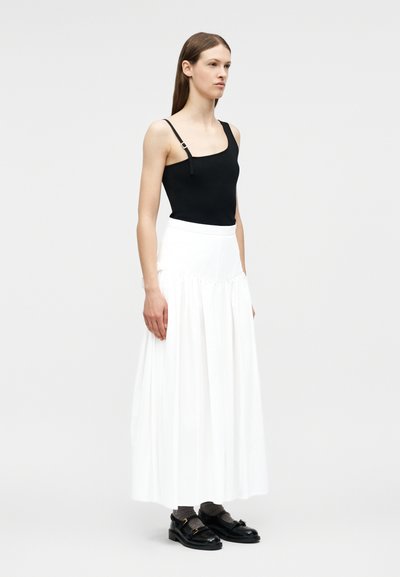 Γυναίκα που στέκεται, φοράει μαύρο ασύμμετρο τοπ με τιράντες, μακριά λευκή φούστα, γκρι κάλτσες και μαύρα παπούτσια σε απλό φωτεινό φόντο.
