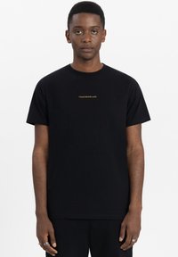 Zwarte katoenen T-shirt met korte mouwen, ronde hals en een klein gouden "TOMORROWLAND" logo op de borst. Eenvoudig, ongecompliceerd ontwerp.