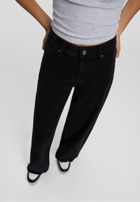 Jean large noir avec poches avant, fermeture à bouton en métal et coutures contrastées. Porté avec des baskets noires et blanches.