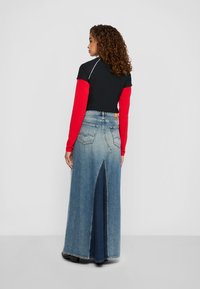 Kvinne med krøllete hår iført svart og rød langermet topp og lang blå denimskjørt med sliss bak, stående mot en enkel lys bakgrunn.
