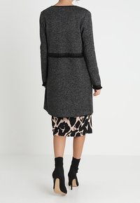Žena má na sobě dlouhý svetr v barvě antracitové šedé přes černo-beige sukni s zvířecím potiskem, doplněný černými vysokými kotníkovými botami.