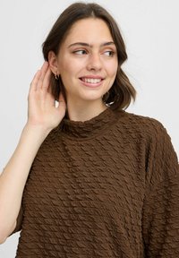 Brauner strukturierter Oberteil mit hohem Kragen, der ein wellenförmiges Muster aufweist. Das Model hat kurzes Haar und trägt schlichte Creolen.