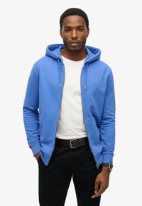 Sudadera azul con cremallera en tejido suave, con capucha, que presenta un bolsillo delantero y detalles en cremallera gris, llevada sobre una camiseta blanca y pantalones negros.