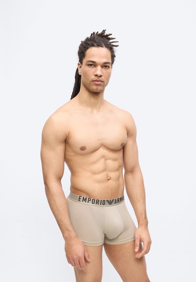 Άνδρας χωρίς πουκάμισο και με κότσους dreadlocks, φορώντας μπεζ εσώρουχα Emporio Armani boxer, στέκεται μπροστά σε μονόχρωμο ανοιχτό γκρι φόντο.