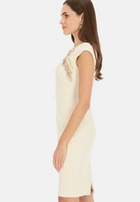 Kremowa dopasowana sukienka z ząbkowanym dekoltem z koronką oraz misternymi zdobieniami na jednym ramieniu. Długość do kolan z rozcięciem po boku.