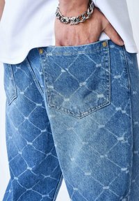 Jean de mezclilla azul claro con un patrón de diamante, que cuenta con un bolsillo trasero y detalles en remaches de cobre. La tela tiene un aspecto desgastado y texturizado.
