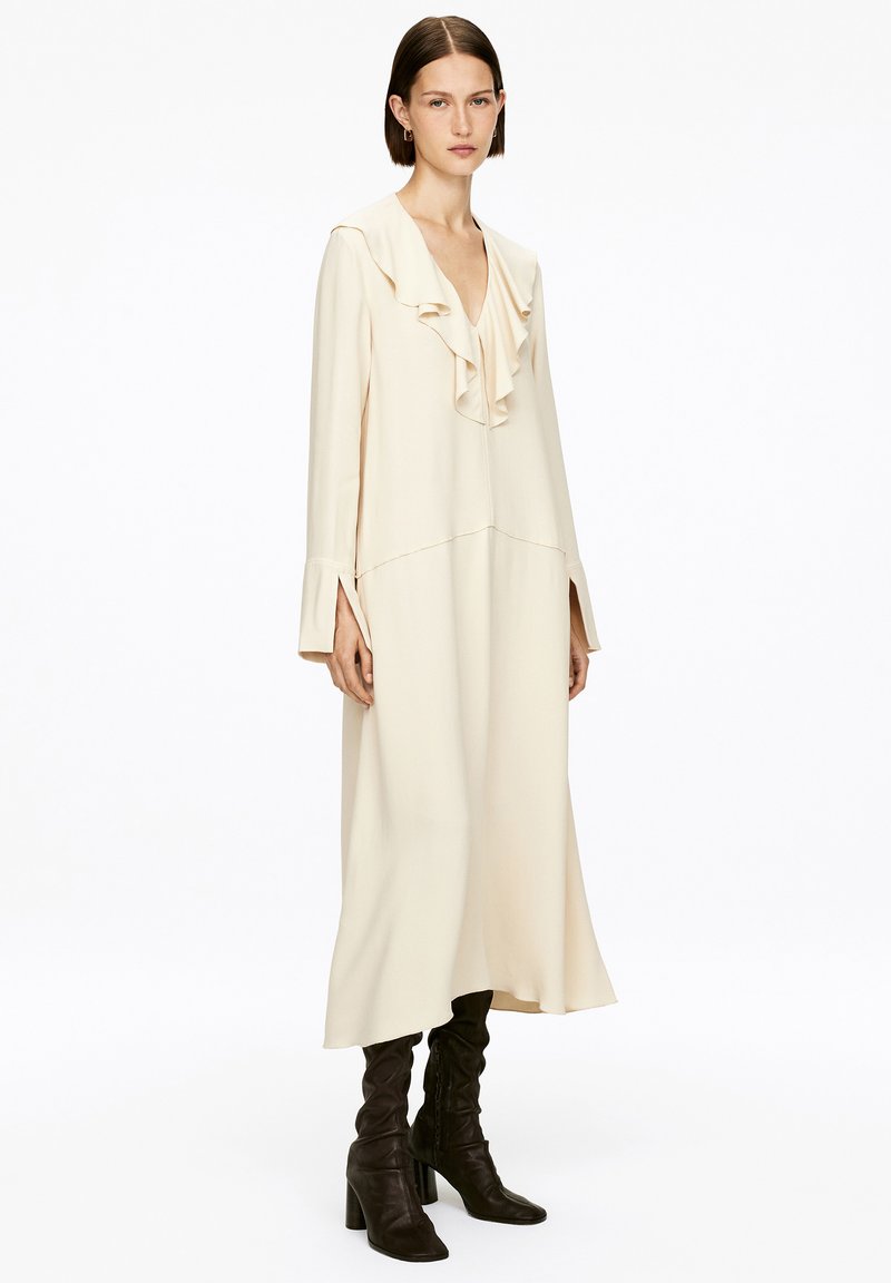 Femme debout, portant une longue robe crème à décolleté en V froncé et à manches longues, associée à des bottes noires à talons hauts jusqu'aux genoux.