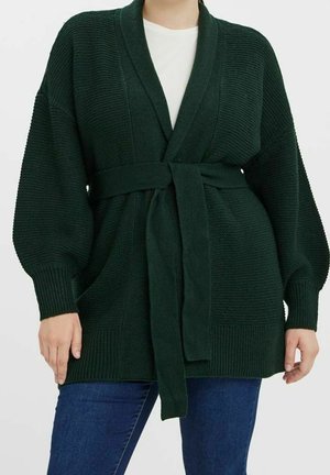 Cardigan en maille vert foncé à manches larges et ceinture nouée à la taille, porté par-dessus un haut blanc avec un jean bleu.