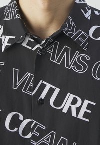 Svart skjorta med knappar framtill som har stora vita textmönster. Materialet ser slätt ut med en klassisk krage och en subtil lyster.