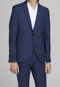 Osoba ubrana w dopasowany granatowy garnitur z dwoma czarnymi guzikami na białej koszulce polo z kołnierzykiem, na jednolitym szarym tle.