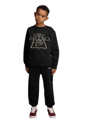 Czarna bluza z beżowym wzorem w klasyczną grę w skakanie, w komplecie z dopasowanymi czarnymi spodniami dresowymi. Model nosi czerwone trampki z białymi akcentami.