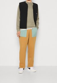 Schwarze gesteppte Weste über einem hellgrünen Langarmshirt, kombiniert mit beigen Jogginghosen mit blau-grünen Taschen. Weiße Sneakers runden das Outfit ab.