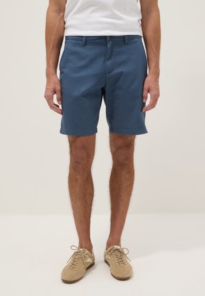 Uomo che indossa pantaloncini blu fino al ginocchio, t-shirt bianca e sneakers beige con lacci, in piedi davanti a uno sfondo uniforme.