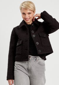 Femme blonde souriante, portant une veste en laine noire à boutons avec des poches sur un haut à pois noirs et un jean gris clair.