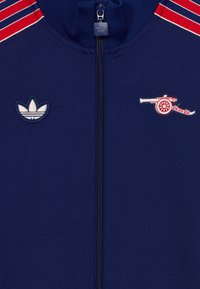 Marinblå zip-up jacka med röda och vita randiga accenter, med broderade Adidas- och Arsenal-kanonlogotyper. Mjuk textur och sportig design.