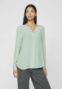 Blusa verde chiaro con scollatura a V e maniche lunghe. Il tessuto è liscio con una leggera lucentezza, presenta un orlo curvo e linee pulite.
