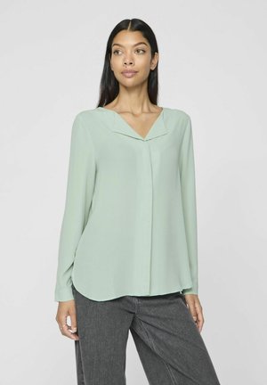 Lys grønn bluse med V-hals og lange ermer. Stoffet er glatt med en lett glans, og har en buet kant og rene linjer.