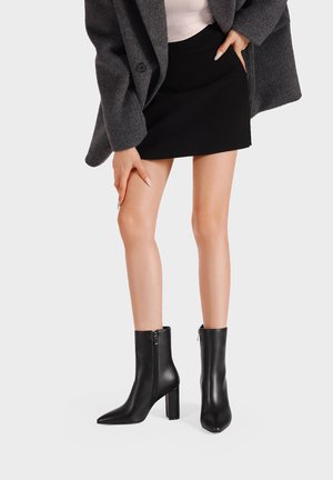 Donna che indossa stivaletti neri con punte appuntite, una minigonna nera, un cappotto grigio e un top bianco, posando con una mano sulla coscia.