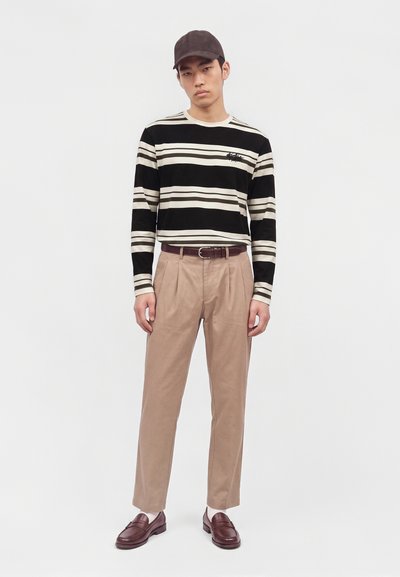 Jeune homme portant une casquette noire, une chemise rayée noire et beige à manches longues, un pantalon beige plissé, une ceinture marron et des mocassins marron avec des chaussettes blanches.