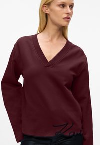 Felpa bordeaux con scollo a V profondo, maniche lunghe e nome del brand ricamato in nero in basso a sinistra. Tessuto morbido e texture.