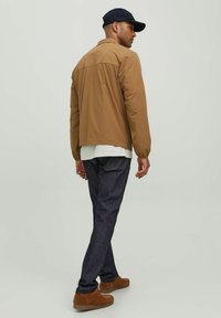 Chaqueta marrón ligera con cuello, combinada con jeans oscuros y zapatos de slip-on marrones, modelo visto de espaldas llevando una gorra azul marino.
