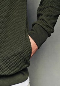 Grøn sweatshirt med struktur og diamantmønster, med ribbede manchetter og en sidelomme. Materialet ser blødt og elastisk ud.